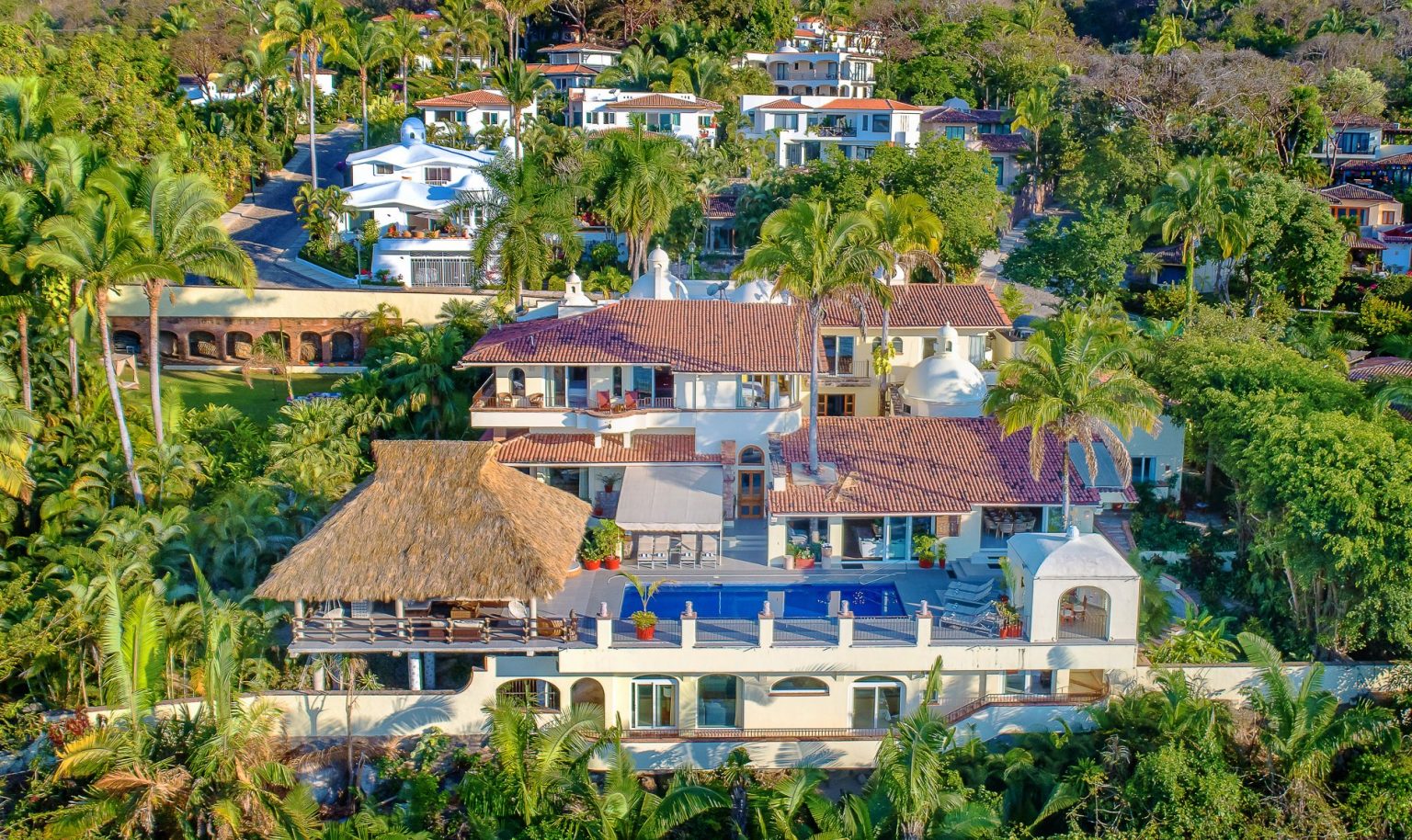 Casa Peregrina Aerial view of a luxury villa complex surrounded by lush greenery and palm trees. The property, part of an exclusive Villa Gallery, features a multi-level villa with red-tiled roofs, an expansive swimming pool, spacious terraces, and a thatched-roof gazebo. Other similar villas are visible in the background. Luxury Villa Retreat